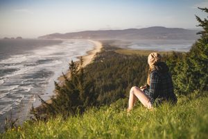 Woman sitting atop grassy hill looking out onto the ocean at a beach