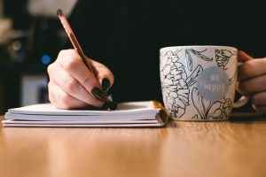 Woman with black nail writing in a journal while drinking from a mug that says Be Happy